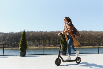 Young beautiful woman in a jacket smiles and rides an electric scooter to work along office buildings