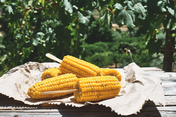 Roasted sweet corn cobs are on the recycled paper on the wooden table in the summer garden
