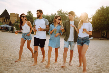 Group of friends cheers and drink beers on the beach. Young  friends sitting together sitting near the sea and enjoy summer party. Beach holiday and summer vacation concept.