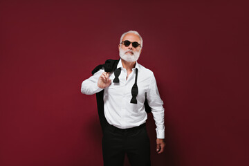 Bearded man in shirt and suit poses on burgundy background. Fashionable guy with white hair in...