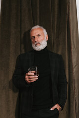 Self confident man in plaid jacket looks into camera and holds whiskey glass. Stylish guy in black clothes posing on velvet background..