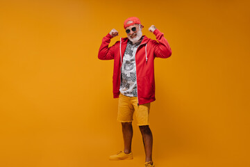 Attractive man in cap, sunglasses and hoodie demonstrates biceps. Cheerful guy with white beard in modern bright clothes posing..