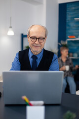 Old woman relaxing on couch holding a cup of coffee and husband is working. Elderly man entrepreneur in home workplace using portable computer sitting at desk while wife is holding tv remote.