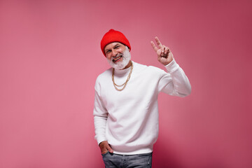 Happy man in orange hat and white sweater showing peace sign on pink background. Adult handsome guy in modern clothes smiling..