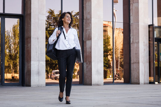 Hispanic Mature Businesswoman With Backpack And Laptop Walking And Laughing In Modern Outdoors