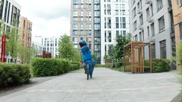 Cheerful dinosaur rides a bicycle among beautiful colored houses. 
A man in a dinosaur costume rides a bicycle among residential areas
