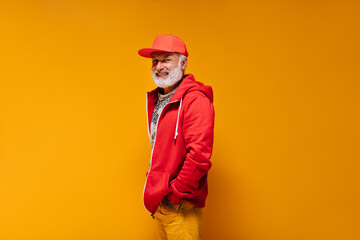 Smiling man in red outfit looks into camera on orange background. Trendy guy with grey beard in bright cap and sweatshirt looking into camera..