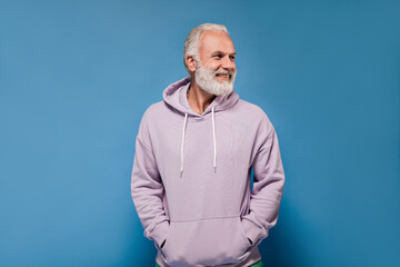 Man in grey hoodie posing on blue background. Cheerful adult guy with white beard in lilac...
