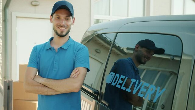 Slow motion portrait of cheerful delivery employee smiling standing near commercial van and parcels and looking at camera. People and job concept.