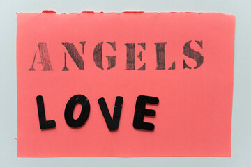 sign with angels written out in stencil font in pencil and love in black chalk letters on coral pink paper