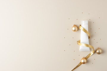 Christmas composition made of cosmetic cream tube, gold ribbon and balls on beige background. Flat lay, copy space.