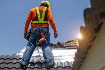 Roof repair, worker with white gloves replacing gray tiles or shingles on house with blue sky as background and copy space, Roofing - construction worker standing on a roof covering it with tiles.
