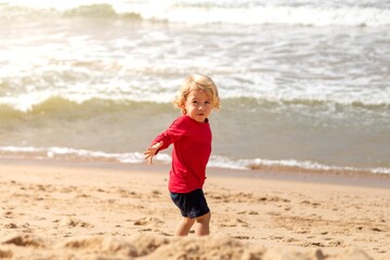 CRIANÇA LOIRA NA PRAIA, ANDANDO EM DIREÇÃO AO MAR. USANDO CAMISA VERMELHA.