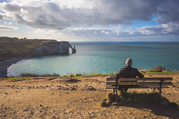 Tourist sitting on a bench admiring the seascape Etretat Aval cliff, rocks and natural arch...