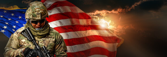 US Army soldier in combat uniforms holding machine gun on the national US flag with gold sunlight...