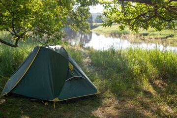 a tourist tent stands on the green grass near the river under a huge oak tree. travel wild in nature