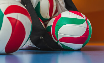 Multiple volleyball balls and a ball bag on school gymnasium floor.