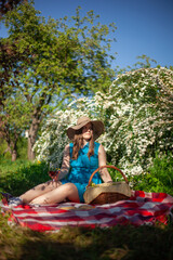 A girl in a hat is resting in a blooming garden with a glass of red wine. Picnic basket, red blanket, book and fruit