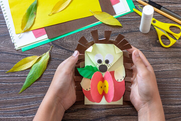 Child is holding backpack of paper greeting card and a postcard hedgehog with an apple on a wooden table. Handmade. Project of children's creativity, handicrafts, crafts for kids. © elena_hramowa