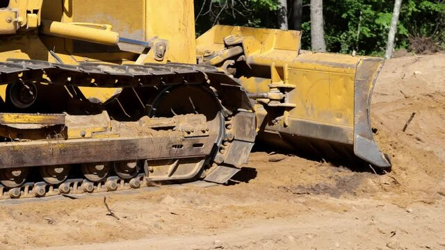 Closeup of blade and track of a working bulldozer as it pushes dirt to level the earth at a new home construction job site. Yellow machine, brown dirt and green trees, with audio.