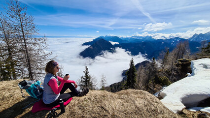 A woman in hiking outfit sipping a tea from thermos with the panoramic view from the top of Alpine peak in Austria. The area is shrouded in thick clouds. A few peaks popping out from the clouds. Happy