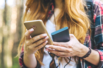 Power bank and smartphone in the hands of a girl with red hair in a shirt in a cage with a black backpack, on the background of a forest road.