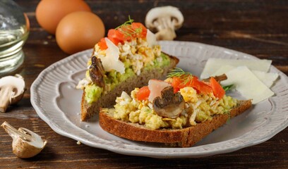 Two toasts with guacamole, scrambled eggs, salmon and mushrooms. On rye bread. Close-up, on a gray plate. Dark wooden background.