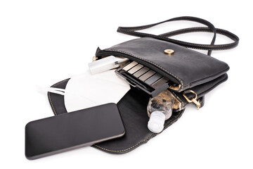Women's accessories spilling out of an open handbag: mobile phone, makeup, lipstick, eye shadows, hand sanitizer and face mask, isolated on white background. Pandemic female essentials concept.
