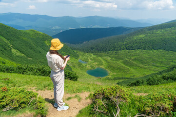 Fototapeta na wymiar Traveler in a hat, resting in nature and using the Internet on a smartphone. A tourist girl photographs a panorama of the mountains and a mountain lake by a mobile phone. Mountain beautiful landscape