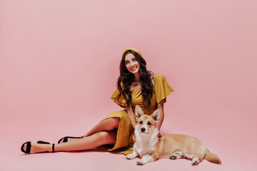 Beautiful girl with long hair in modern yellow dress and dark shoes smiling and posing with corgi on pink isolated background..