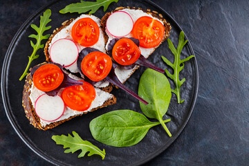 Healthy toasts with cream cheese, cherry tomatoes and spinach on black plate. Bruschetta with cream cheese and fresh herbs on a dark background. Copy space. Top view