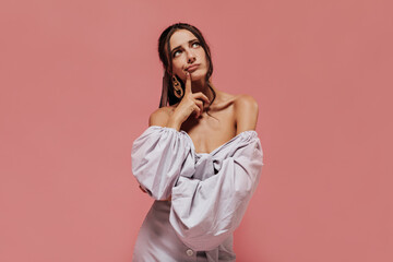 Pensive slim lady with tanned skin and brunette hair in gold accessories in wide sleeve lilac shirt looking up and posing..