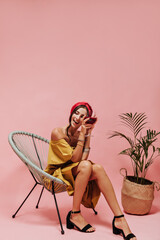 Lovely modern woman in good mood with stylish earrings, bright outfit and heels sitting on chair, smiling and posing with glass..