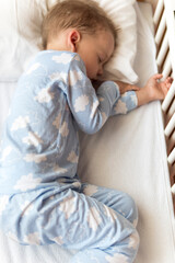 top view Cute little 2-3 years preschool baby boy kid sleeping sweetly in white crib during lunch rest time in blue pajama with pillow at home. Childhood, leisure, comfort, medicine, health concept
