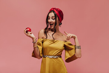 Surprised wavy haired trendy lady with cool hairstyle in red bandana and bright sundress looking at apple on isolated pink backdrop..