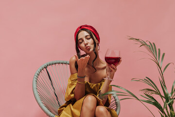 Curly haired pretty lady in red headband and yellow trendy sundress blowing kiss and holding glass on isolated backdrop..