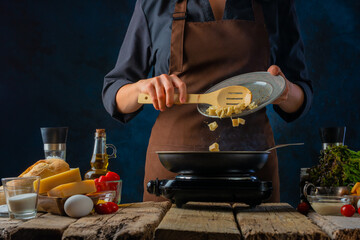 There are many ingredients for making Caesar salad on a wooden table. A chef in a dark uniform is frying chicken in a skillet for a salad. Bright paint ingredients and dark background.
