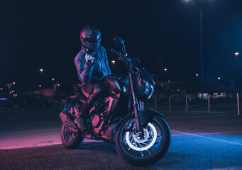 Naklejka na ściany i meble Motorcyclist sits on a motorbike in neon light in an empty parking lot at night