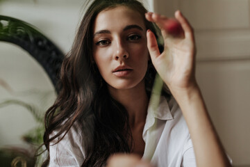 Fototapeta premium Charming modern girl with tanned skin and curly brunette hairstyle in stylish white shirt looking into camera in apartment..