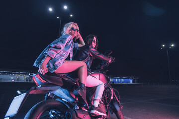 Fototapeta na wymiar a couple of girls in love on a motorbike in the rays of neon light in an empty parking lot at night
