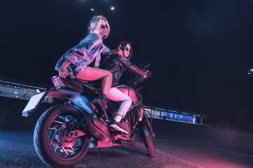 Fototapeta na wymiar a couple of girls in love on a motorbike in the rays of neon light in an empty parking lot at night