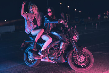 Plakat a couple of girls in love on a motorbike in the rays of neon light in an empty parking lot at night