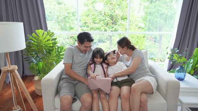 Asian parents with little daughter and son sitting on sofa in living room using digital tablet play games or watch movie together. Happy family having fun weekend activity with technology at home.