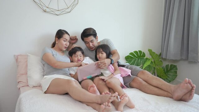 Asian parents with little daughter and son sitting on the bed in bedroom using digital tablet play games or watch movie together. Happy family having fun weekend activity with technology at home.