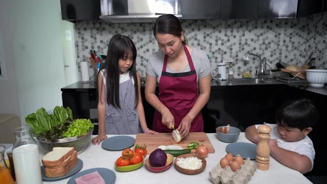 Asian mother teach little daughter and son making vegetable and ham sandwich on the table for breakfast in kitchen. Mom and child boy and girl kids enjoy weekend activity cooking together at home.