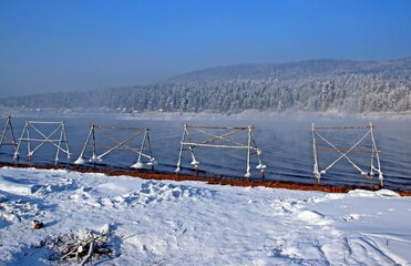 A guardrail of the shoreline of the river. Icy steel structures stand in the water. A haze over the water and a coniferous forest on the opposite bank.