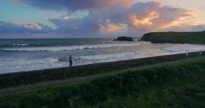 Aerial: Man walking along ocean wall on stormy day with rough ocean. People are camping on a farm. Annestown Beach, Tramore, Ireland