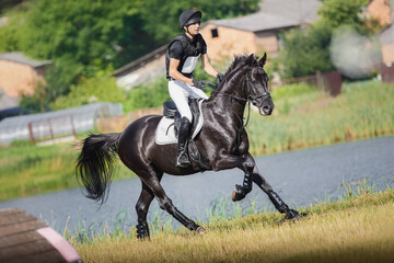 portrait of rider man and black stallion horse galloping fast near lake during eventing cross...