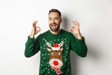 Christmas, holidays and celebration. Happy young man in green sweater, winking and showing okay...