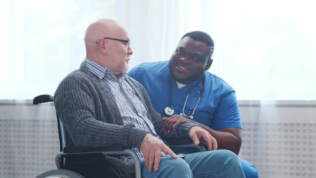 African-American caregiver and old disabled man in a wheelchair. Professional nurse and handicapped patient in a nursing home. Assistance, rehabilitation and health care.